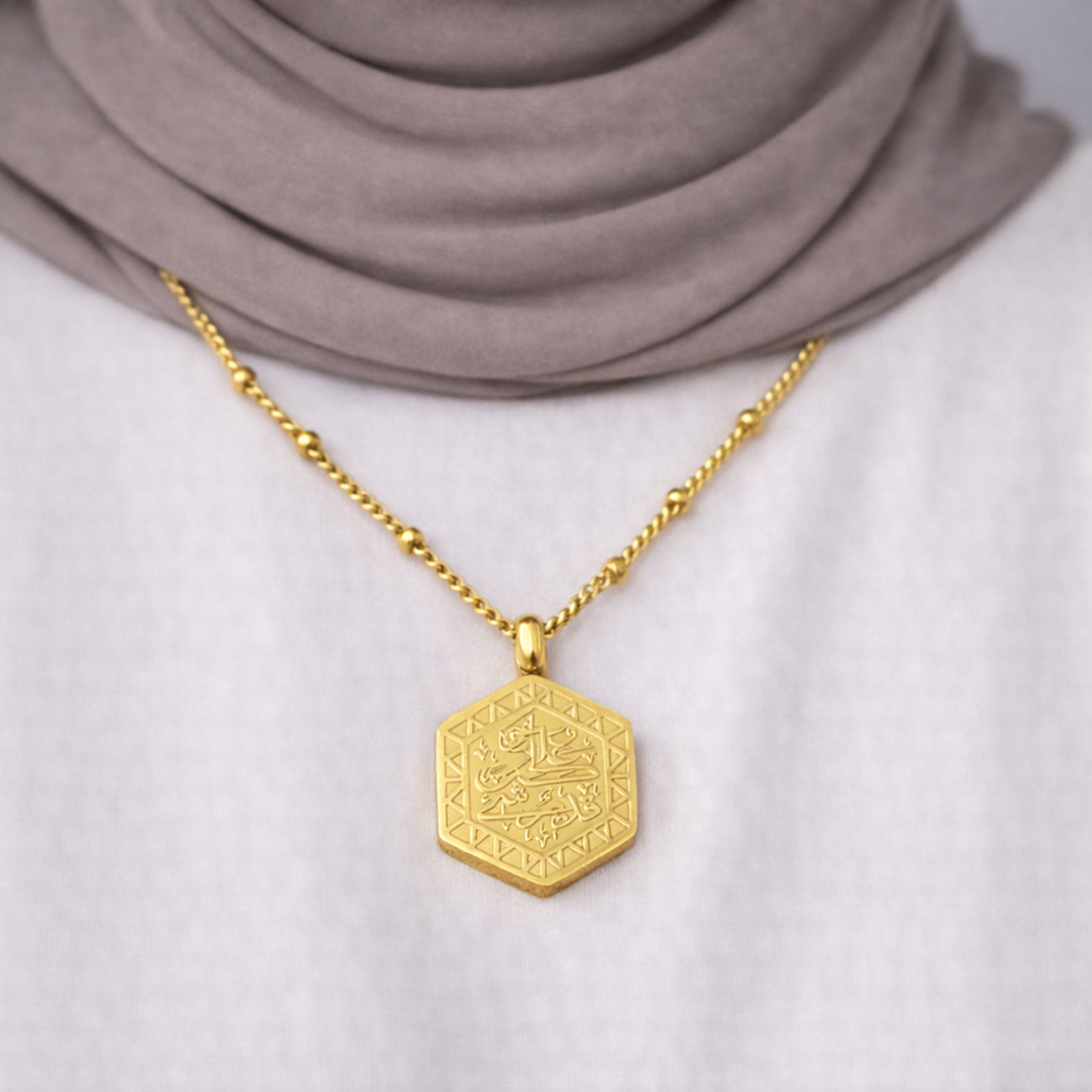 Gold Al-Qadīr Necklace worn by a modest woman, featuring Quran verse meaning “And He is capable of all things,” styled with a white top and neutral hijab.